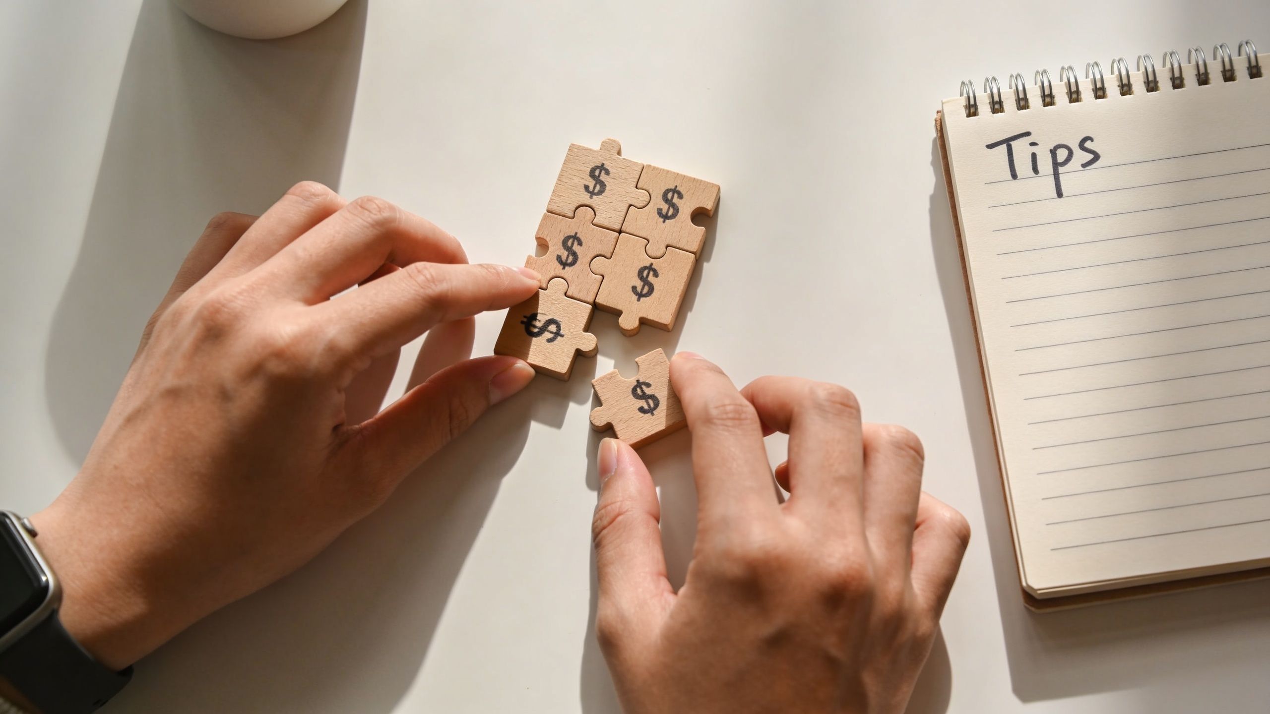 A pair of hands connecting wooden puzzle pieces with dollar signs next to a notebook labeled Tips.